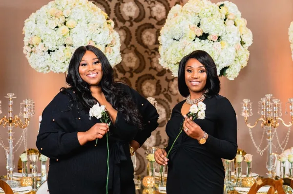 Two women in black dresses holding white roses standing in front of a floral and event design display.