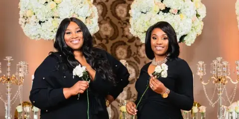 Two women in black dresses holding white roses standing in front of a floral and event design display.