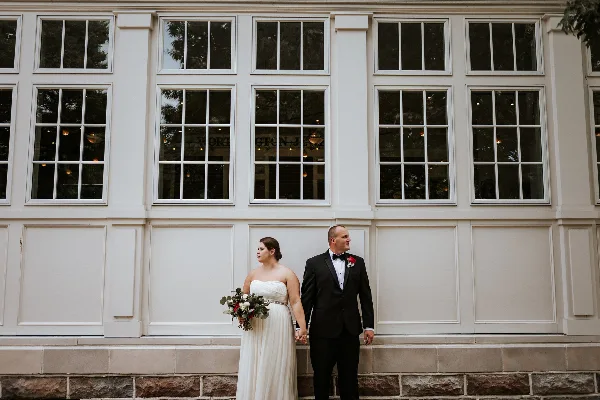 Bride and groom holding hands outside a white wedding venue, with the bride carrying a bouquet.