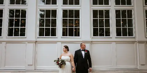 Bride and groom holding hands outside a white wedding venue, with the bride carrying a bouquet.
