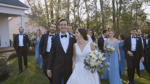 A bride and groom walking with their wedding party outdoors on a lawn.