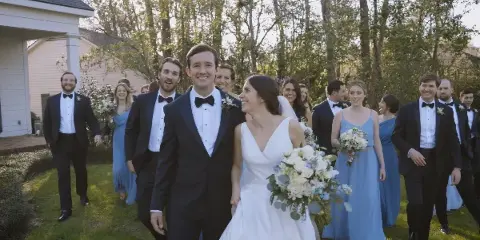 A bride and groom walking with their wedding party outdoors on a lawn.