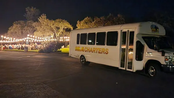 A white MLA Charters shuttle bus parked at an outdoor event venue at night with string lights and guests in the background.