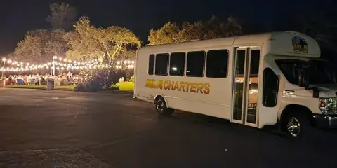 A white MLA Charters shuttle bus parked at an outdoor event venue at night with string lights and guests in the background.