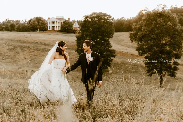 Bride and groom walking through a grassy field with a white manor house visible in the background.