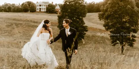 Bride and groom walking through a grassy field with a white manor house visible in the background.