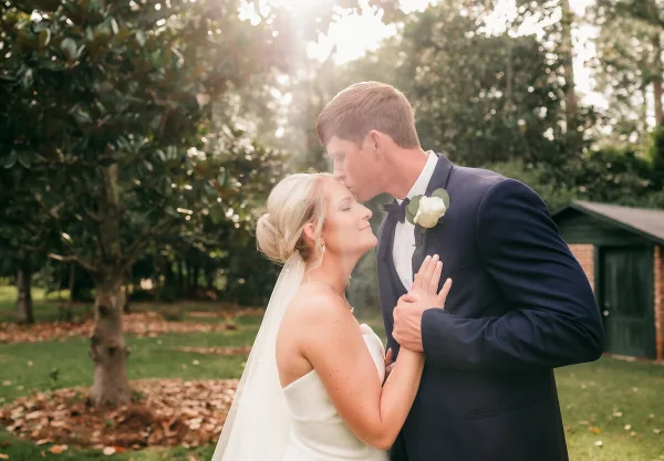 A bride and groom sharing a tender moment outdoors, with the groom kissing the bride's forehead.