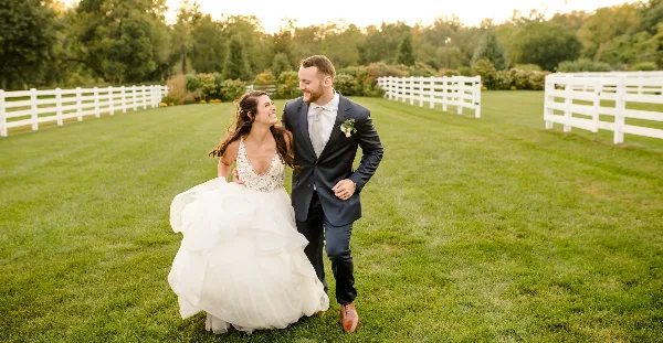 A bride and groom walking together on a grassy lawn with white fences in the background.