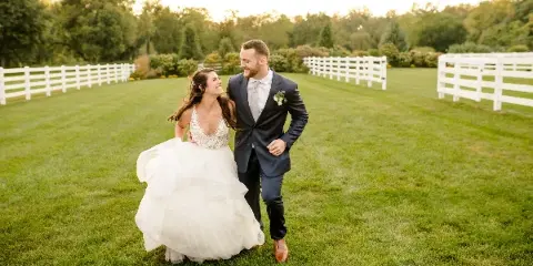 A bride and groom walking together on a grassy lawn with white fences in the background.