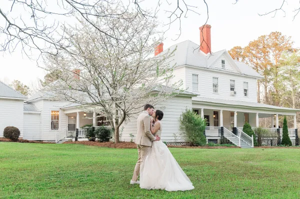 A bride and groom kissing on the lawn in front of a white historic estate house with a wraparound porch.