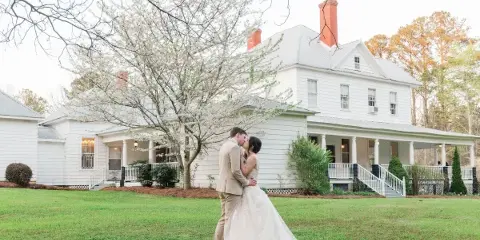 A bride and groom kissing on the lawn in front of a white historic estate house with a wraparound porch.