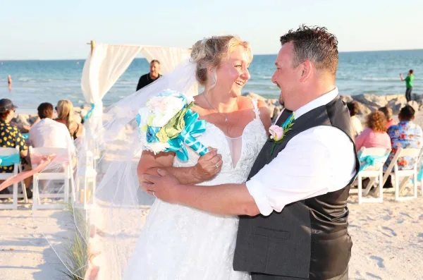 A bride and groom embracing during a beach wedding ceremony with a white arch and guests in the background.