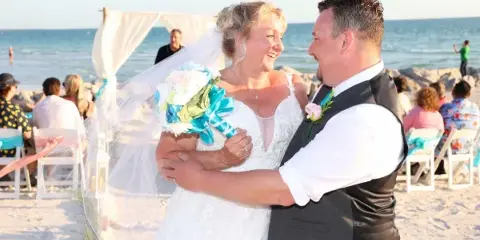 A bride and groom embracing during a beach wedding ceremony with a white arch and guests in the background.