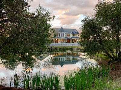 A Victorian farmhouse reflected in a pond surrounded by trees and greenery.