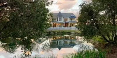 A Victorian farmhouse reflected in a pond surrounded by trees and greenery.