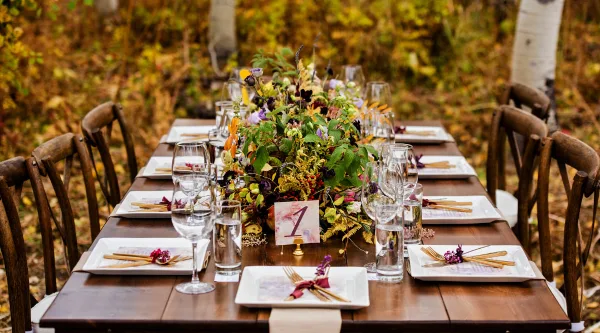 A long wooden event table set with white plates, glassware, and a large floral centerpiece, surrounded by wooden chairs in an outdoor setting.
