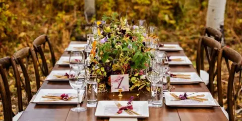 A long wooden event table set with white plates, glassware, and a large floral centerpiece, surrounded by wooden chairs in an outdoor setting.
