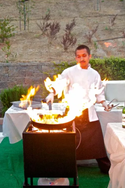 A chef in a white uniform cooking with a large flame in a wok at an outdoor event.