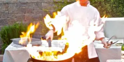 A chef in a white uniform cooking with a large flame in a wok at an outdoor event.