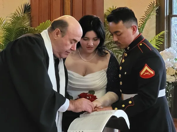 A wedding officiant in clerical robes performing a ring exchange ceremony with a bride and groom.