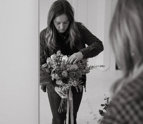 A woman in a plaid shirt arranging a wedding bouquet with roses and greenery.