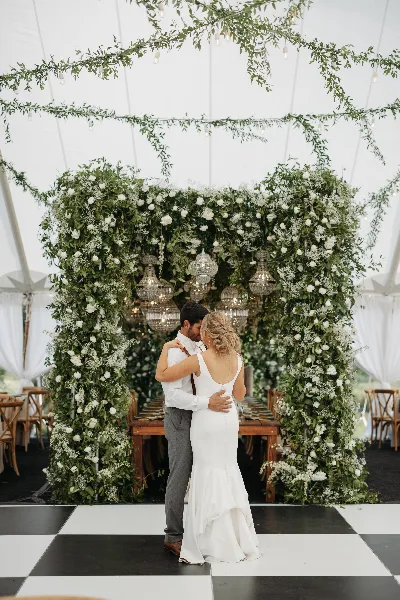 A bride and groom embracing in front of a lush floral wall and chandeliers at a wedding reception.