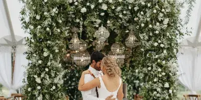 A bride and groom embracing in front of a lush floral wall and chandeliers at a wedding reception.