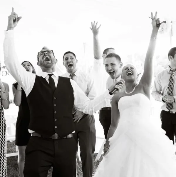 A DJ holding a microphone and a bride dancing with raised hands at a wedding reception.
