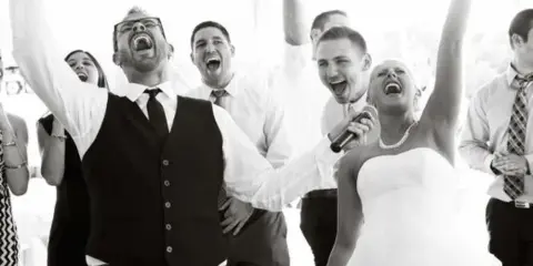 A DJ holding a microphone and a bride dancing with raised hands at a wedding reception.