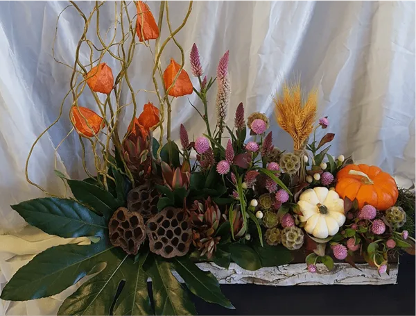A professional floral arrangement featuring orange and white pumpkins, wheat stalks, and mixed flowers in a rustic white container.