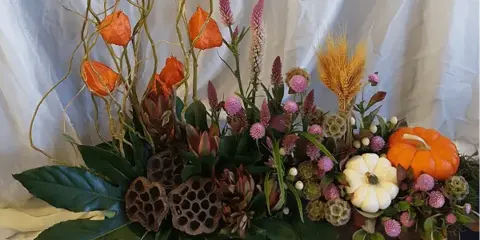 A professional floral arrangement featuring orange and white pumpkins, wheat stalks, and mixed flowers in a rustic white container.