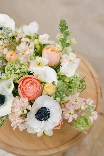 A close-up of a floral arrangement with white anemones, peach roses, and green hydrangeas resting on a wooden surface.