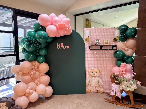 A custom event backdrop with a dark green arch, pink and green balloon garlands, and a personalized name sign.