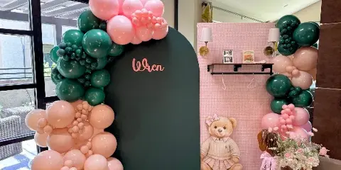 A custom event backdrop with a dark green arch, pink and green balloon garlands, and a personalized name sign.