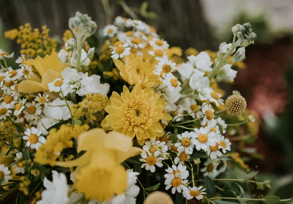 A close-up photograph of a wedding floral arrangement featuring yellow chrysanthemums, white daisies, and greenery.