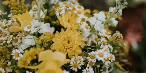 A close-up photograph of a wedding floral arrangement featuring yellow chrysanthemums, white daisies, and greenery.