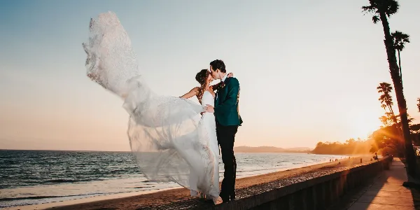 A bride and groom kissing on a beach boardwalk at sunset with the bride's veil blowing in the wind.