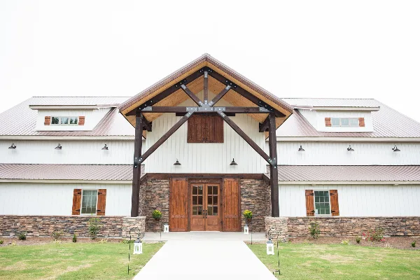 Exterior view of a white barn with a stone base and wooden double doors, featuring a large timber frame entrance and metal roof.