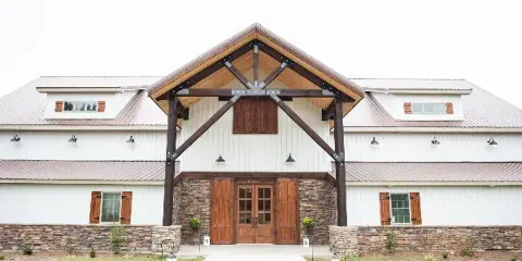 Exterior view of a white barn with a stone base and wooden double doors, featuring a large timber frame entrance and metal roof.
