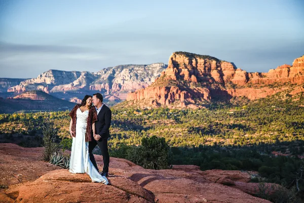 A bride and groom standing on a red rock ledge with a mountainous landscape in the background.