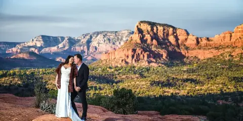 A bride and groom standing on a red rock ledge with a mountainous landscape in the background.
