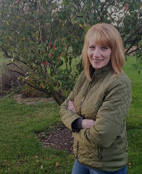 Portrait of a smiling woman with blonde hair wearing a green quilted jacket, standing outdoors in a garden setting.