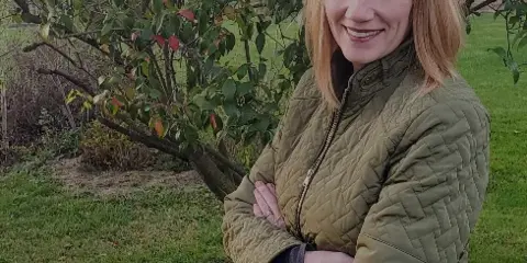 Portrait of a smiling woman with blonde hair wearing a green quilted jacket, standing outdoors in a garden setting.