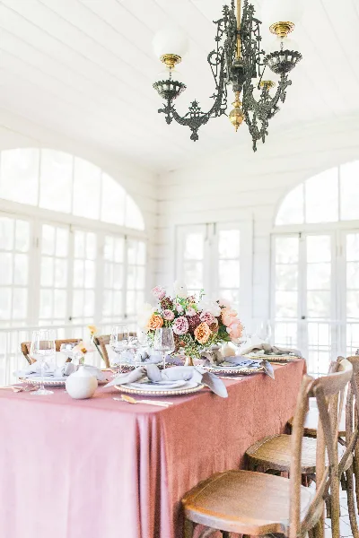 A professionally styled wedding reception table featuring a floral centerpiece, gold-rimmed plates, and a pink tablecloth in a bright venue.