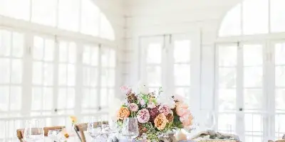 A professionally styled wedding reception table featuring a floral centerpiece, gold-rimmed plates, and a pink tablecloth in a bright venue.
