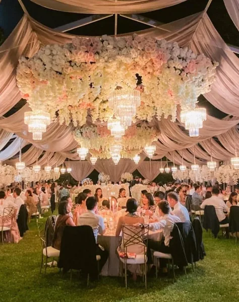 An outdoor wedding reception venue featuring a large floral and chandelier installation suspended from a draped ceiling, with guests seated at round tables on a lawn.