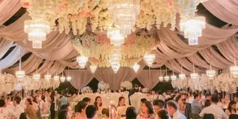 An outdoor wedding reception venue featuring a large floral and chandelier installation suspended from a draped ceiling, with guests seated at round tables on a lawn.