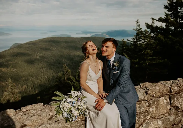 A bride and groom laughing together on a stone wall with a scenic mountain and water view in the background.