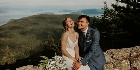 A bride and groom laughing together on a stone wall with a scenic mountain and water view in the background.