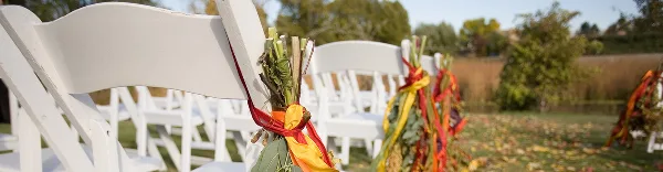 Rows of white folding chairs decorated with floral arrangements and ribbons set up outdoors for a wedding.